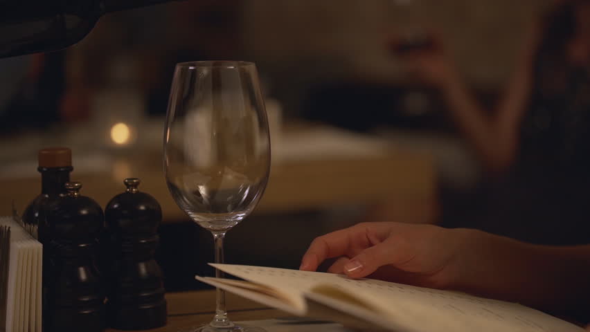 Close up footage of wine pouring from bottle to glass while Caucasian female looking at menu sitting at restaurant in slowmotion