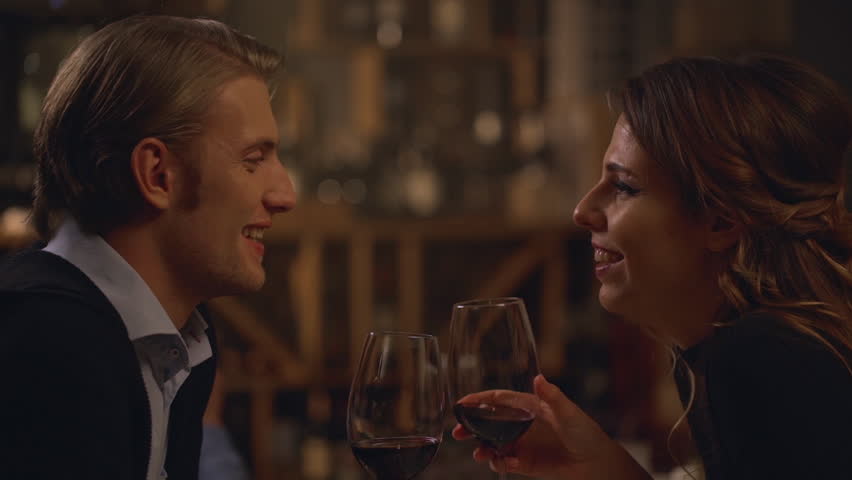 Beautiful Caucasian male and female in love sitting at restaurant drinking wine from glasses and kissing each other in slowmotion