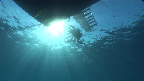 scuba divers ascending back to the boat with the sun and surface and boat ladder background underwater