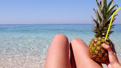 Sexy female legs on a beach with crystal clear turquoise water in the background. Young woman holding pineapple cocktail and enjoying summer vacation