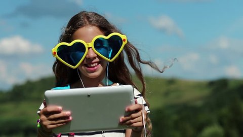 Attractive girl talks via tablet computer. Merry girl in big sunglasses in the shape of hearts communicates via tablet pc. Female in glasses with white tablet pc. Girl communicates through skype