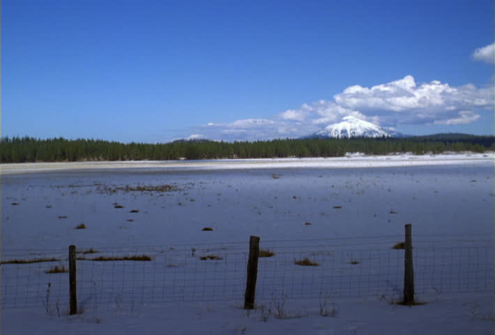Right pan over snow-covered pasture to ranch gate with barn and snowy peaks in distance