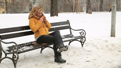 Portrait of a blond woman in warm clothes sitting on a park bench suffering from cold blowing her nose with tissue outdoors in winter.  She has a flue and fever.
