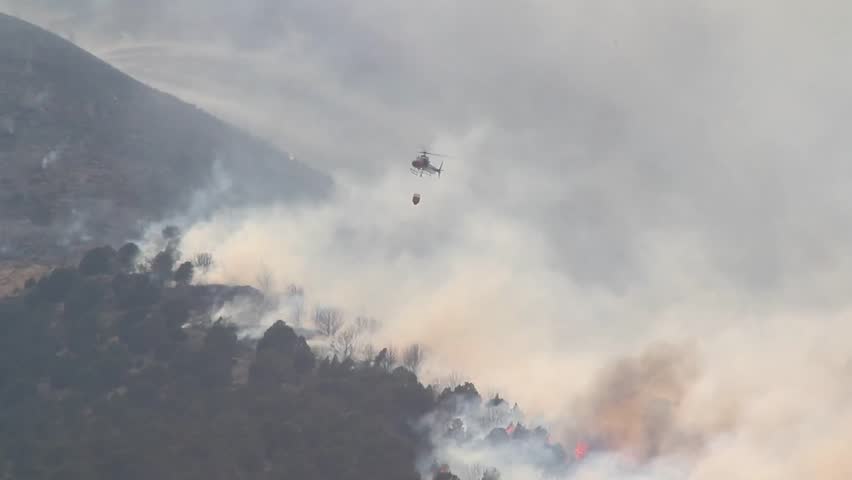 A helicopter battles a gigantic wildfire on a dry mountainside, dropping