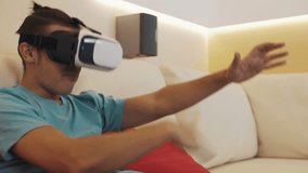 Young caucasian joyful man wears virtual reality headset in blue shirt with hair knot sitting on white leather sofa in modern apartment living room describing his experience.