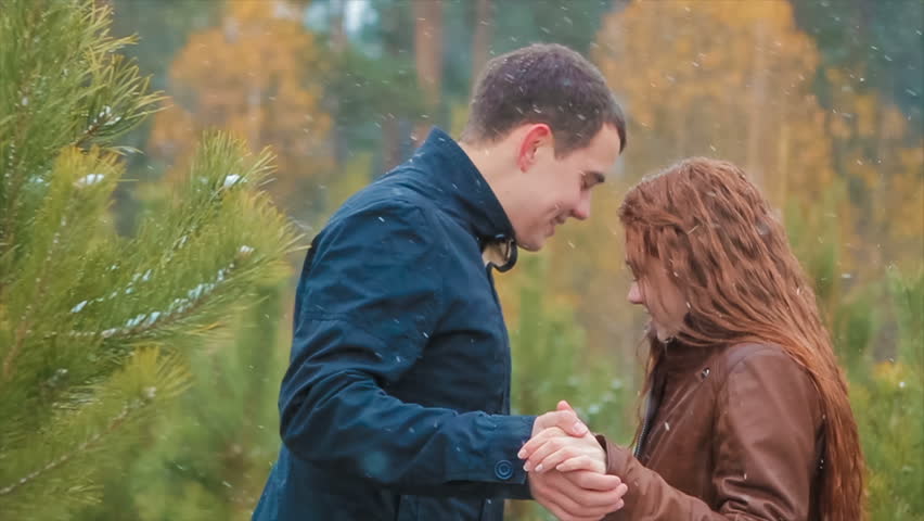 Red Hair Woman and Brunette Man Holding Hands and Interrupted by Phone Call. Man is Taking His phone and Letting go of Woman's Hand. They in Pine Forest. it in Green,yellow,orange Colours With Snow