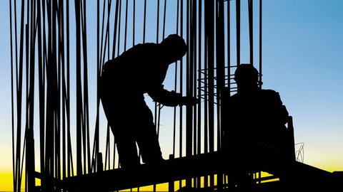 Behind a metal frame workers secure poles with wire to the base of a reinforced concrete pillar on a construction site at sunset