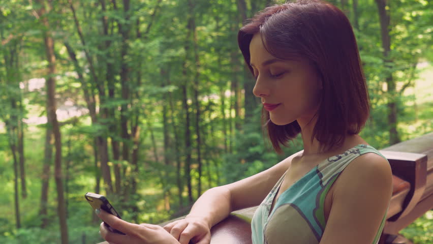 A young beautiful lady is sitting and holding a smartphone in her hands. She is looking on the screen and typing a message or surfing the internet. 
