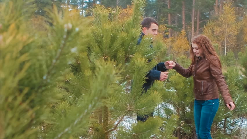 Dark-Hair Young Man and Woman With Long Curly Red Hair Are in Pine Forest.they Holding Hands While Having Fun and Laughing. It's Winter, It's Snowing. Pine Branch in Foreground.