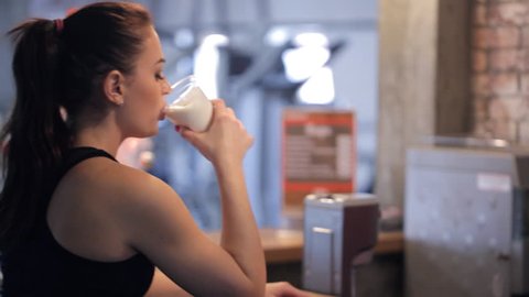 Barman in fitness center give protein shake to young slim beautiful woman. In sports center redheaded lady with ponytail in black sportswear sitting at wooden bar, drink nutrition milk cocktail as
