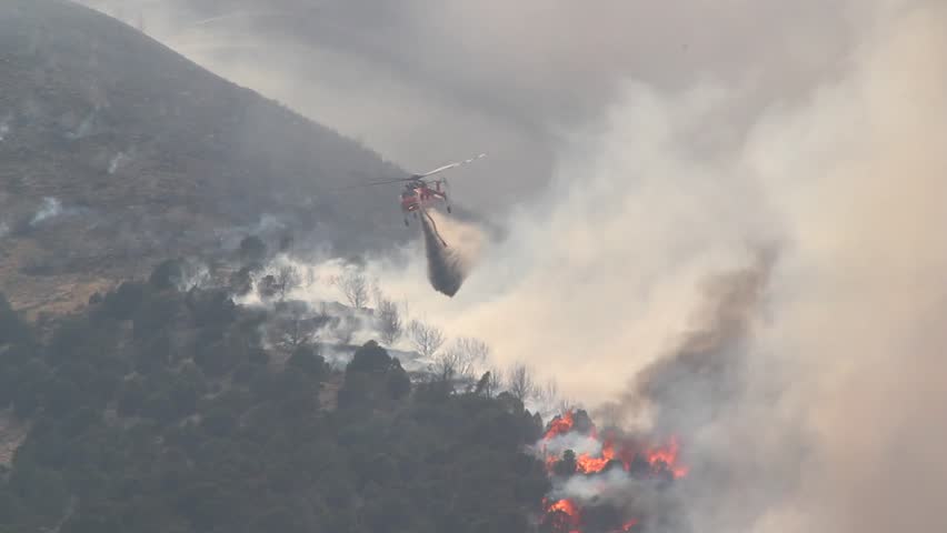 A helicopter battles a gigantic wildfire on a dry mountainside, dropping