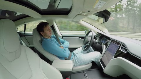 Male driver sleeping behind the self-driving steering wheel of an autonomous autopilot driverless car. Man fell deeply asleep while driving along the countryside road in luxury all-electric vehicle