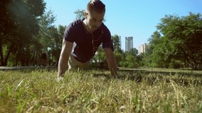 A handsome man in trendy outfit makes push ups during the video. He is wearing a blue T-shirt and an armband for the smartphone. The male is working out in the park with an urban background.