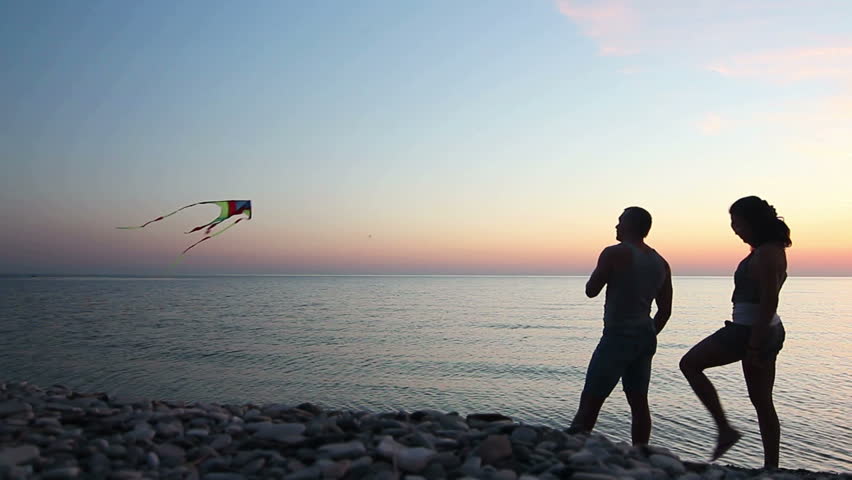 Man flies a kite on the shore of the sea at sunset, couple embracing  on beach a
