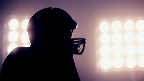 CU Silhouette of male American football player putting on his protective helmet against bright stadium illumination lights. Bearded man. 50 FPS 4K UHD RAW edited footage