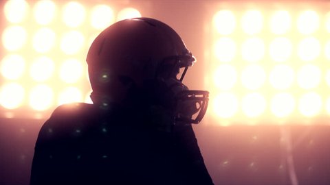 CU Silhouette of male American football player putting on his protective helmet against bright stadium illumination lights. Bearded man. 50 FPS 4K UHD RAW edited footage