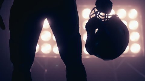 Silhouette of male American football player walking towards camera against bright stadium illumination lights. Bearded man. 4K UHD RAW edited footage