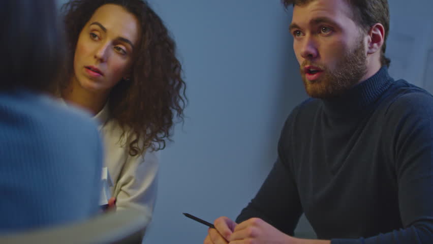 Panning shot of curly designer and her colleague in glasses looking at mobile phone and laughing about something while chatting in office lit by sunlight beaming through window blinds 