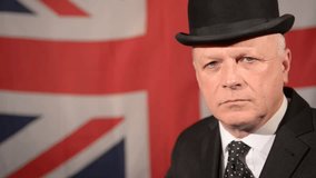 Unhappy British businessman wearing traditional black bowler hat and matching suit, gives a thumbs down hand gesture and keeps it in frame, against a large Union Jack background in a light breeze.