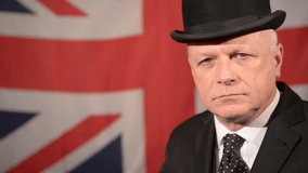 Unhappy British businessman wearing traditional black bowler hat and matching suit, gives a thumbs down hand gesture then lowers his hand, stands against large Union Jack background in a light breeze.