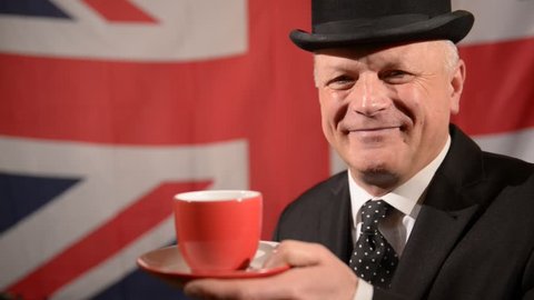 Smiling British businessman wearing traditional black bowler hat and matching suit, sipping/drinking tea then raises eyebrows in approval, stands against large Union Jack background in a light breeze.