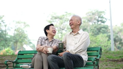 Slow motion happy Asian senior elder couple sitting and drinking tea, coffee in morning park. Having conversation and showing love