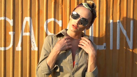 Closeup of sensual blond woman in light brown coverall, bandana and sunglasses posing near gas station booth - video in slow motion