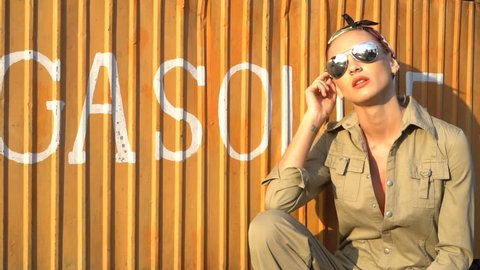 Closeup of sensual blond woman in light brown coverall, bandana and sunglasses posing near gas station booth - video in slow motion