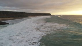 Aerial footage of waves breaking on rocky shore against cloudy sky. As the waves crash they create texture from white sea foam. Scenic view of ocean and mountain cliffs during sunset.  