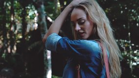Closeup of stunning blond woman in blue denim shirt with backpack looking into the camera and smiling while standing on the bridge over tropical forest background - video in slow motion