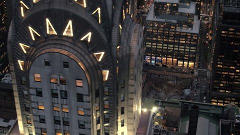 AERIAL HELI SHOT: Flying past lit with lights iconic Chrysler Building rising above modern office buildings and busy crowded New York streets after the sunset. Industry machinery in construction pit