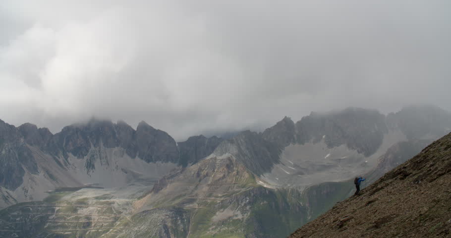 Man hiking up steep mountains with mountain range in background and storm clouds building.