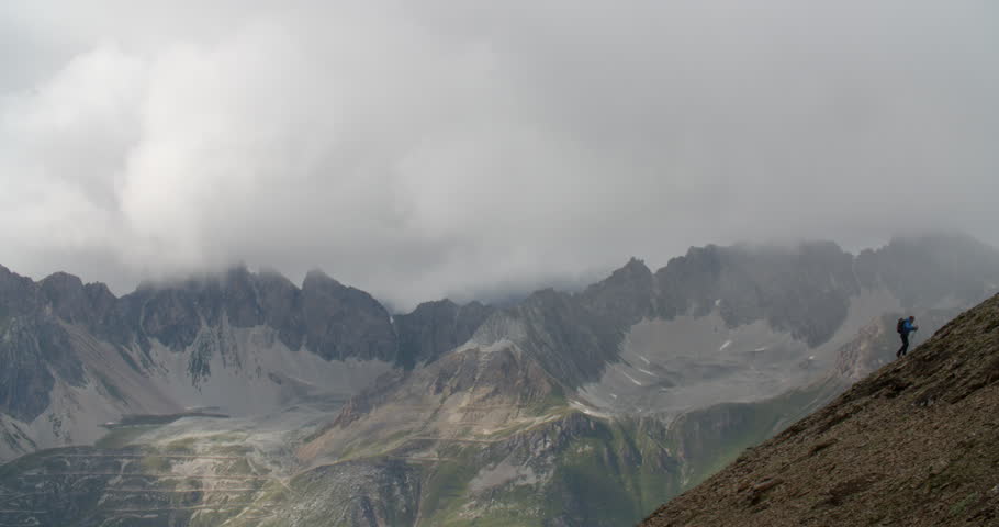 Man hiking up steep mountains with mountain range in background and storm clouds building.