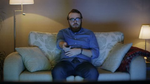 Portrait Shot of a Man Sitting on a Sofa in His Living Room, Watching TV, Changing Channels. Floor Lamps are Turned ON. Shot on RED EPIC-W 8K Helium Cinema Camera.
