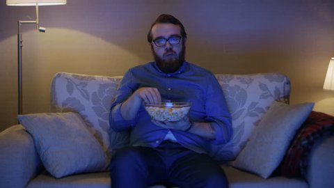 Portrait Shot of a Man Sitting on a Sofa in His Living Room, Eating Popcorn and Watching TV. Floor Lamps are Turned ON. Shot on RED EPIC-W 8K Helium Cinema Camera.