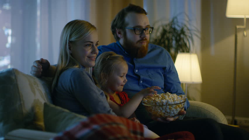 Father, Mother and Little Girl Watching TV. They Sit on a Sofa in Their Cozy Living Room and Eat Popcorn. It's Evening. Shot on RED EPIC-W 8K Helium Cinema Camera. Royalty-Free Stock Footage #24653873