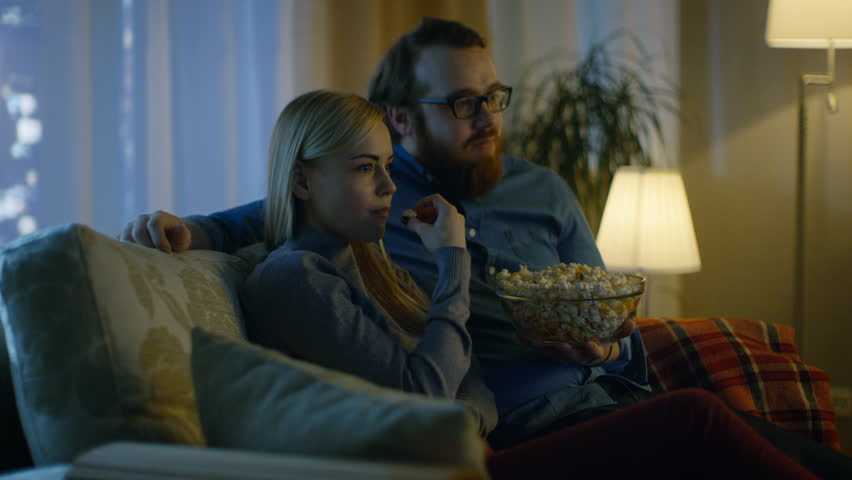 Couple Watching TV. They Sit on a Sofa in Their Cozy Living Room and Eat Popcorn. It's Evening. Shot on RED EPIC-W 8K Helium Cinema Camera. Royalty-Free Stock Footage #24653885