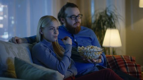 Couple Watching TV. They Sit on a Sofa in Their Cozy Living Room and Eat Popcorn. It's Evening. Shot on RED EPIC-W 8K Helium Cinema Camera.