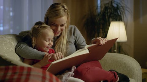 Mother and Her Cute Daughter are Sitting On a Couch in the Living Room. They're Reading Children's Book. It's Evening. Shot on RED EPIC-W 8K Helium Cinema Camera.