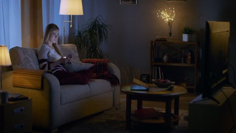 In the Evening Beautiful Young Woman Relaxes on a Couch in Her Cozy Living Room. She Uses Her Smartphone and Simultaneously Watches TV.  Shot on RED EPIC-W 8K Helium Cinema Camera.