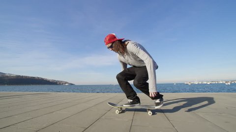 CLOSE UP SLOW MOTION: Young pro skateboarder skateboarding and jumping ollie flip trick on promenade along the coast in sunny summer. Skateboarder jumping kickflip trick with skateboard near the ocean
