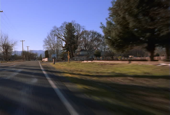 Hood-mount POV of car traveling on rural road past farmhouse