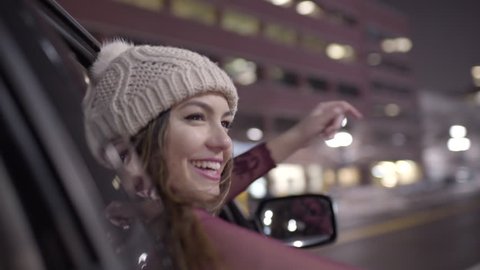 Carefree Young Woman Leans Out Car Window At Night In Downtown, She Waves To People, Raises Her Arms In The Air With Excitement 