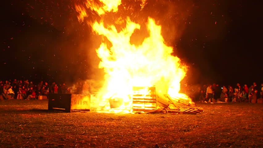 Unrecognizable people watch as huge bonfire burns at night.