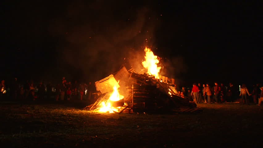 Unrecognizable people watch as huge bonfire gets lit and burns at night, time