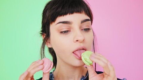 Young and beautiful girl with nude make up isolated on fresh spring backdrop, licks two macarons in her hands to taste it and make a choice what to bite first, closeup portrait