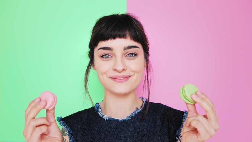 Attractive smiling young brunette isolated on splitted pistachio and rose red background holds two tasty cookies in her hands and makes her choice, then bite left macaron in pastel pink color Royalty-Free Stock Footage #24691346