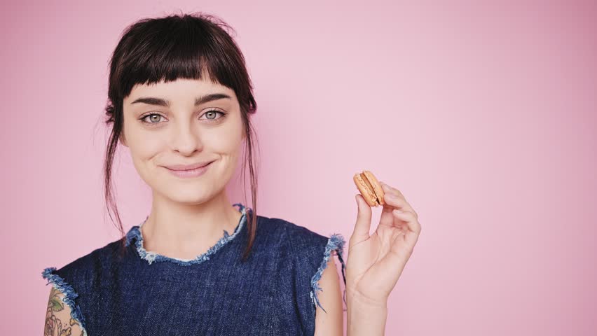 Young fresh and beautiful brunette girl isolated on side on pale pink background smiles, thinks about something, looks on small macaron cookie in her hand and bites it, wearing denim dress