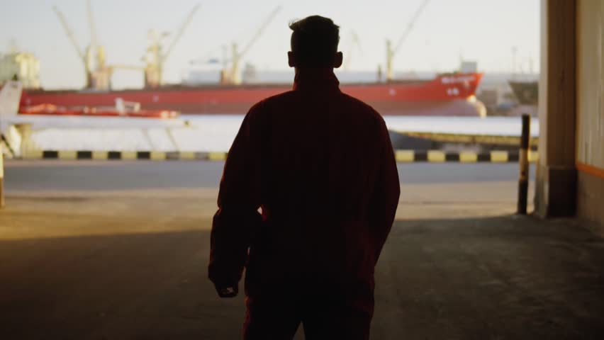 Silhouette of a worker in orange uniform walking through the harbour storage by the sea during his break and raising his hands up. Happy young man.