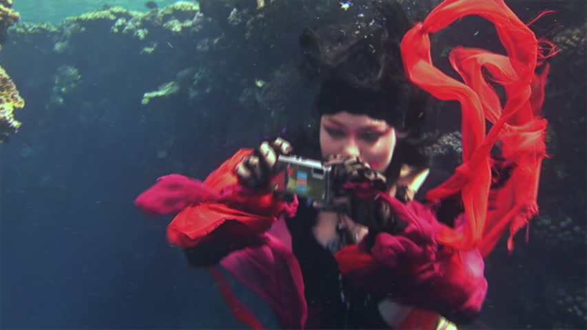 Young girl model free diver underwater in red costume of pirate in Red Sea. Filming a movie. Actress smiling at camera. Extreme sport in marine landscape, coral reefs, ocean inhabitants.